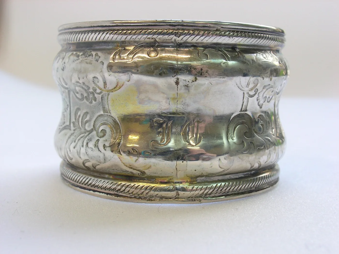 Napkin ring with repoussé scrollwork and a monogram, showing warm pink-gold patches where the silver plating has worn through along the roped rims