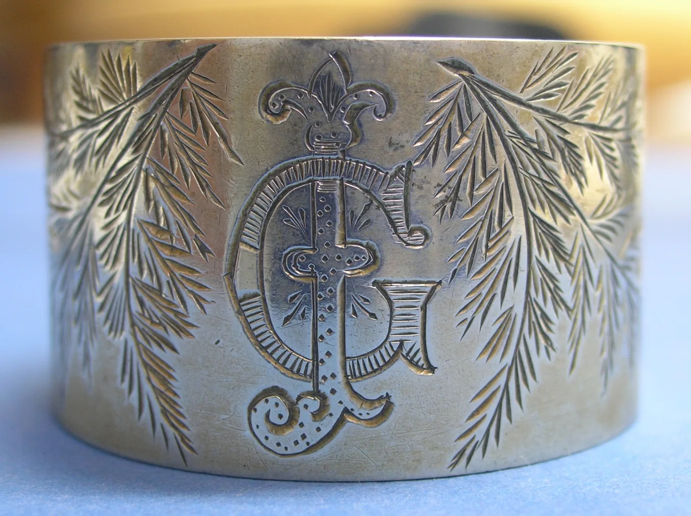Close view of an elaborate interlaced monogram cut into a silver napkin ring, flanked by engraved fern fronds