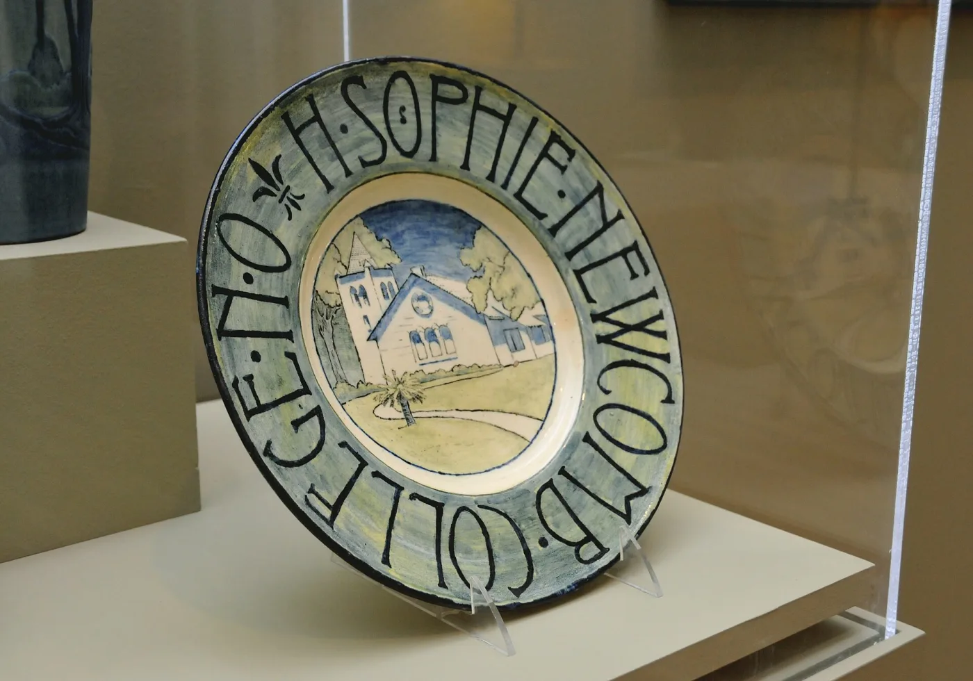 Display case holding a circular pottery plate painted in blue and cream with a building among trees, ringed by hand-lettered capitals reading H. Sophie Newcomb College