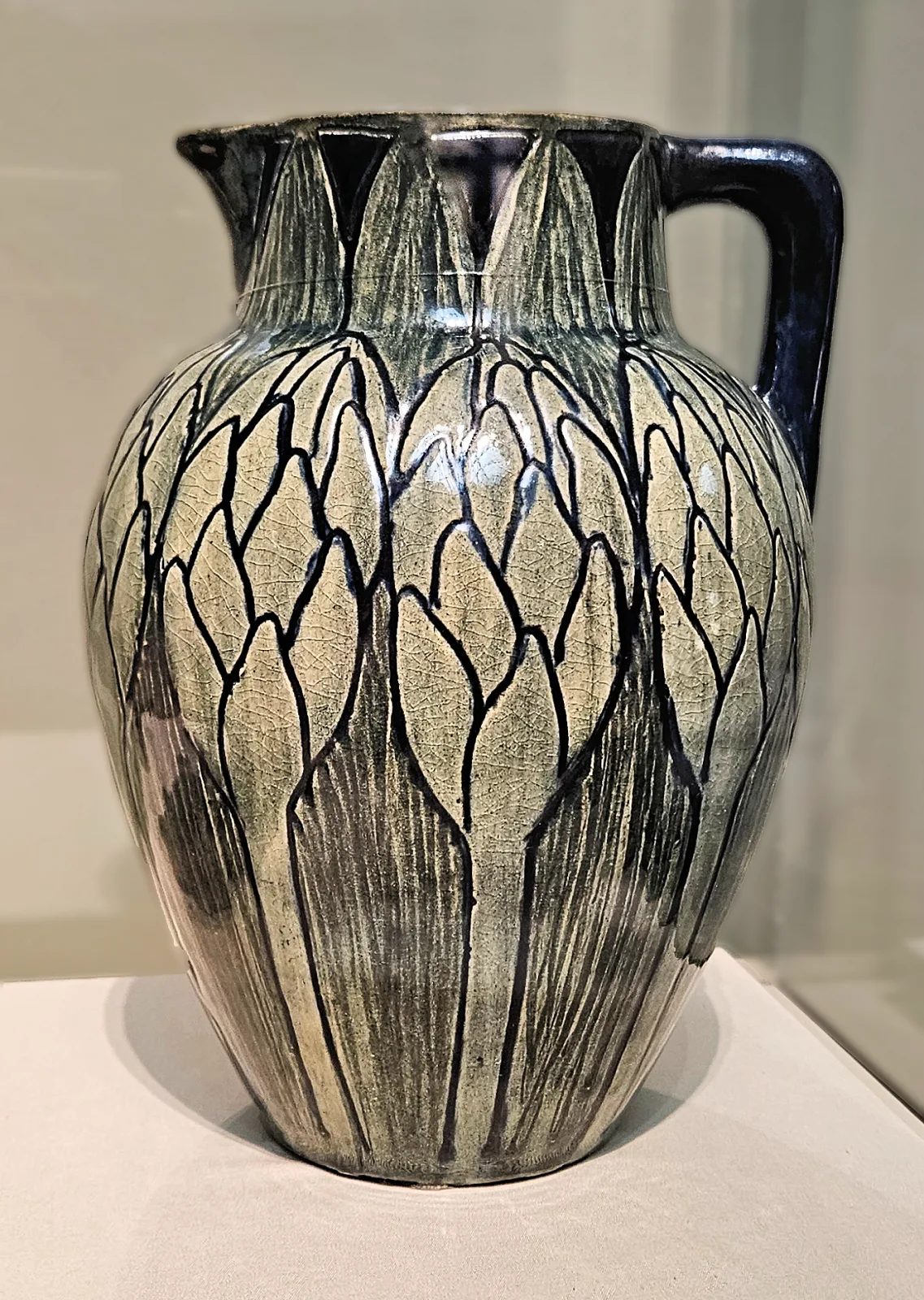 Two-handled pottery jug in glossy grey-blue, its body covered with long pointed leaves outlined in dark blue-black, the glaze thick and reflective