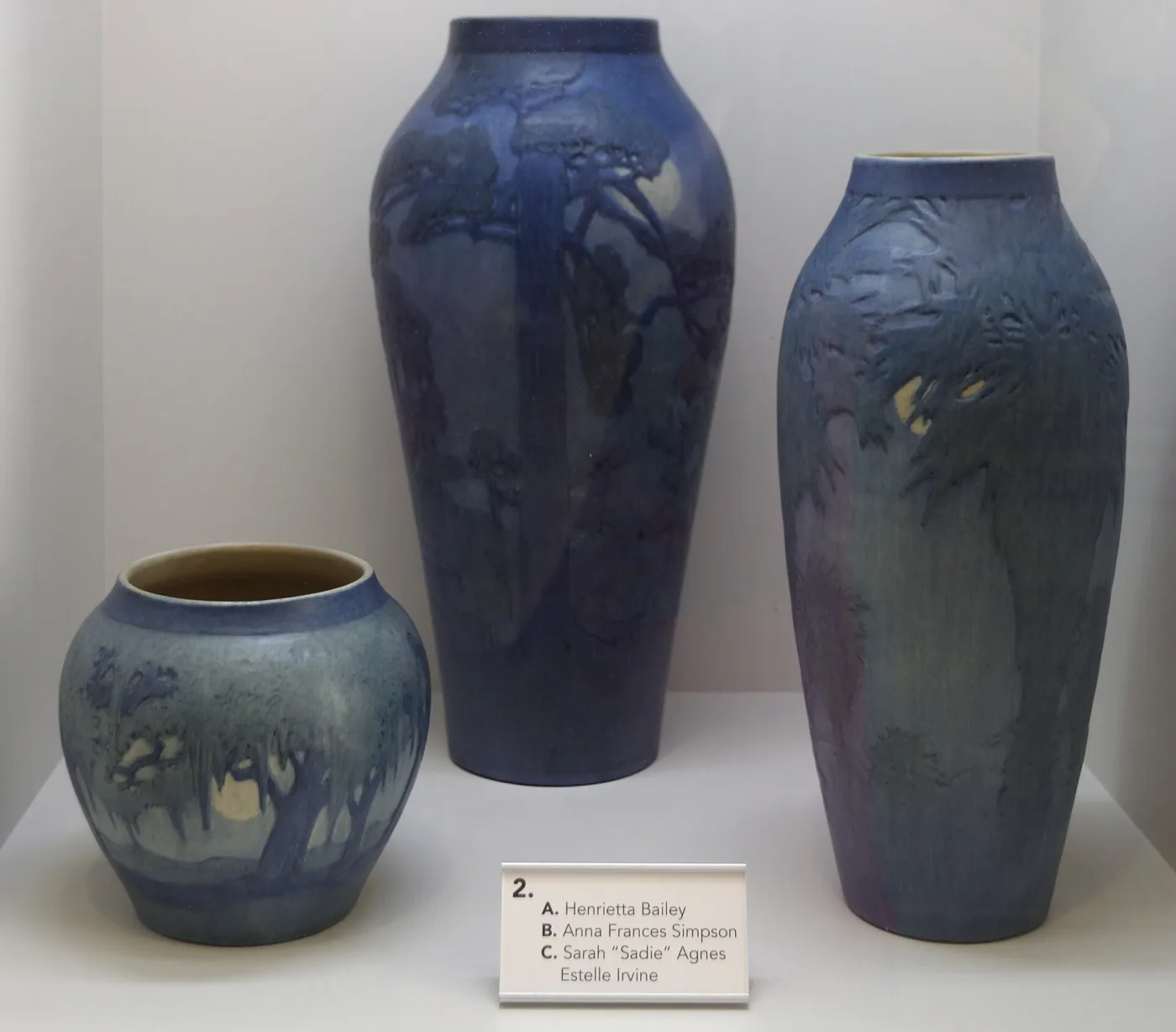 Museum case holding three blue matte vases of different sizes carved with trees and landscapes, with a printed label below naming three decorators