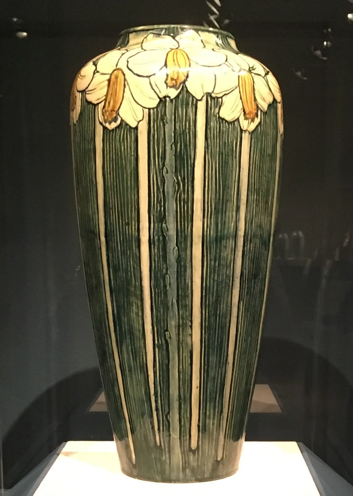 Tall vase lit from above in a dark case, its lower body deeply carved into vertical ribs in green and cream, the shoulder ringed with pale yellow daffodil heads