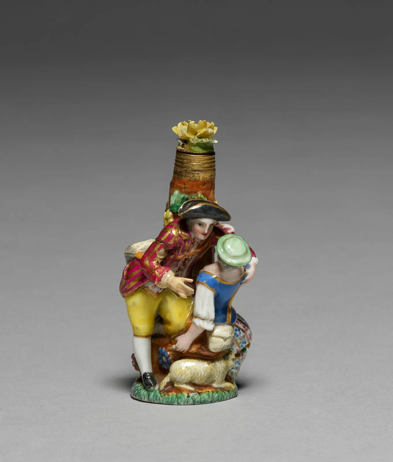 Porcelain scent bottle modelled as a shepherd in a pink coat and a shepherdess in a blue bodice seated with a lamb, the bottle neck rising behind them with a gilt metal collar and a moulded yellow flower stopper