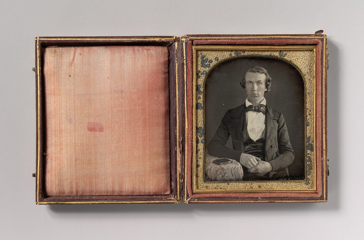 Small hinged case lying open: a plain pink silk pad fills the left half, and on the right a portrait of a seated young man sits behind glass under a shaped gilt mat