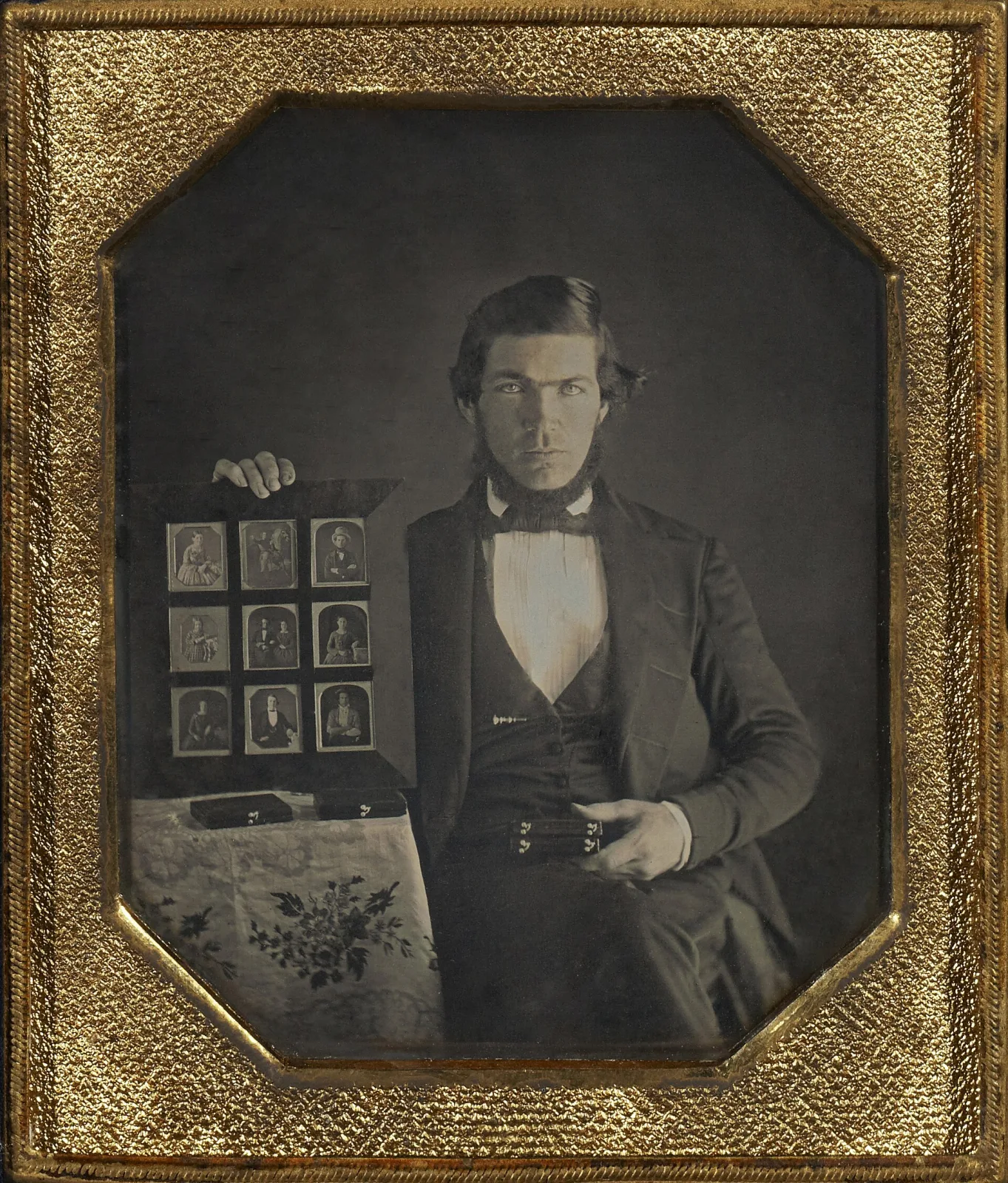 Daguerreotype portrait in an ornate gilt frame of a standing man in a dark coat, one hand resting on a table and the other holding an open case displaying a grid of small portrait plates