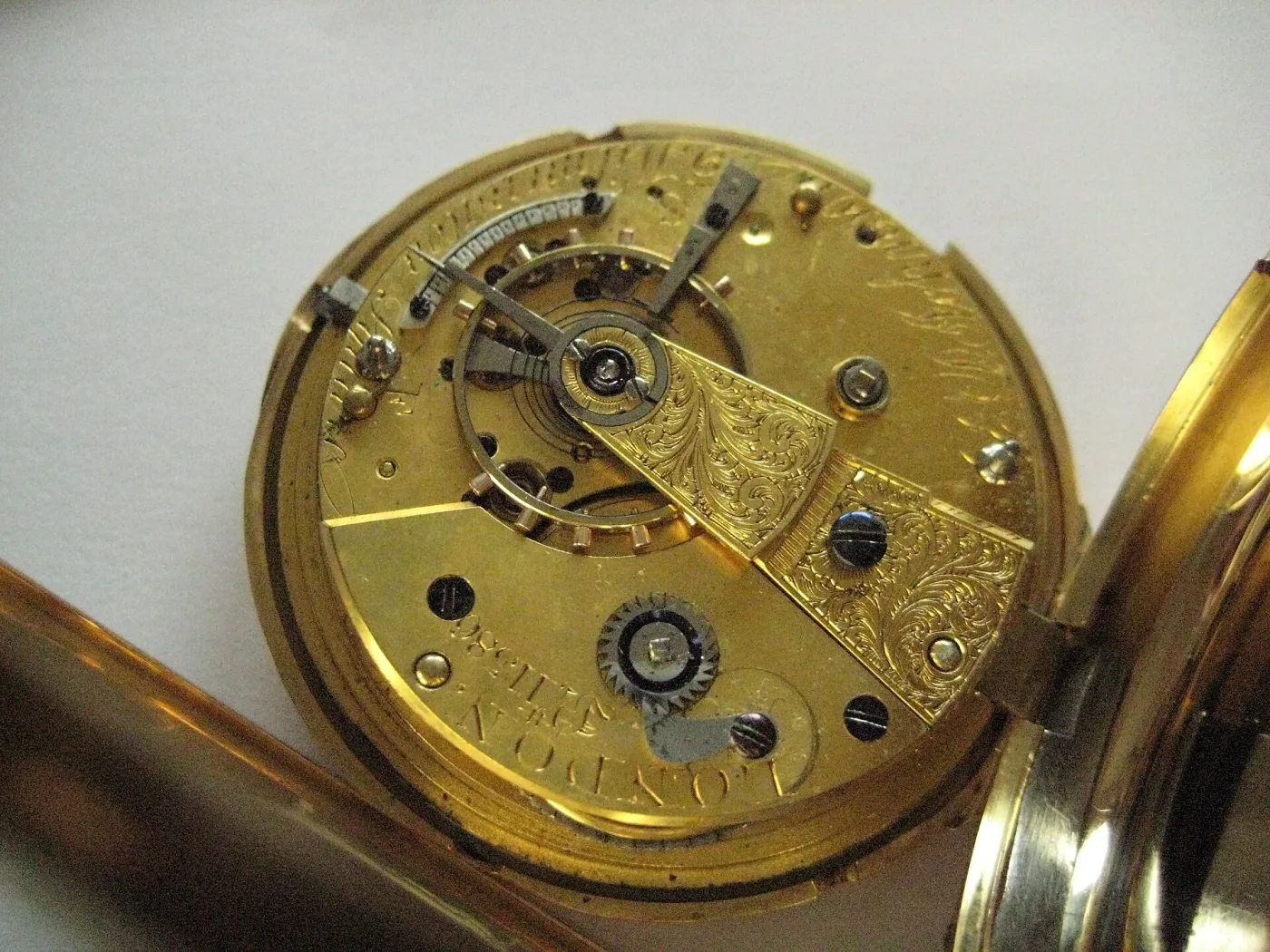 Gilt watch movement lying in its opened case, seen from the back, with an engraved balance cock over the balance wheel and the maker's name engraved across the plate