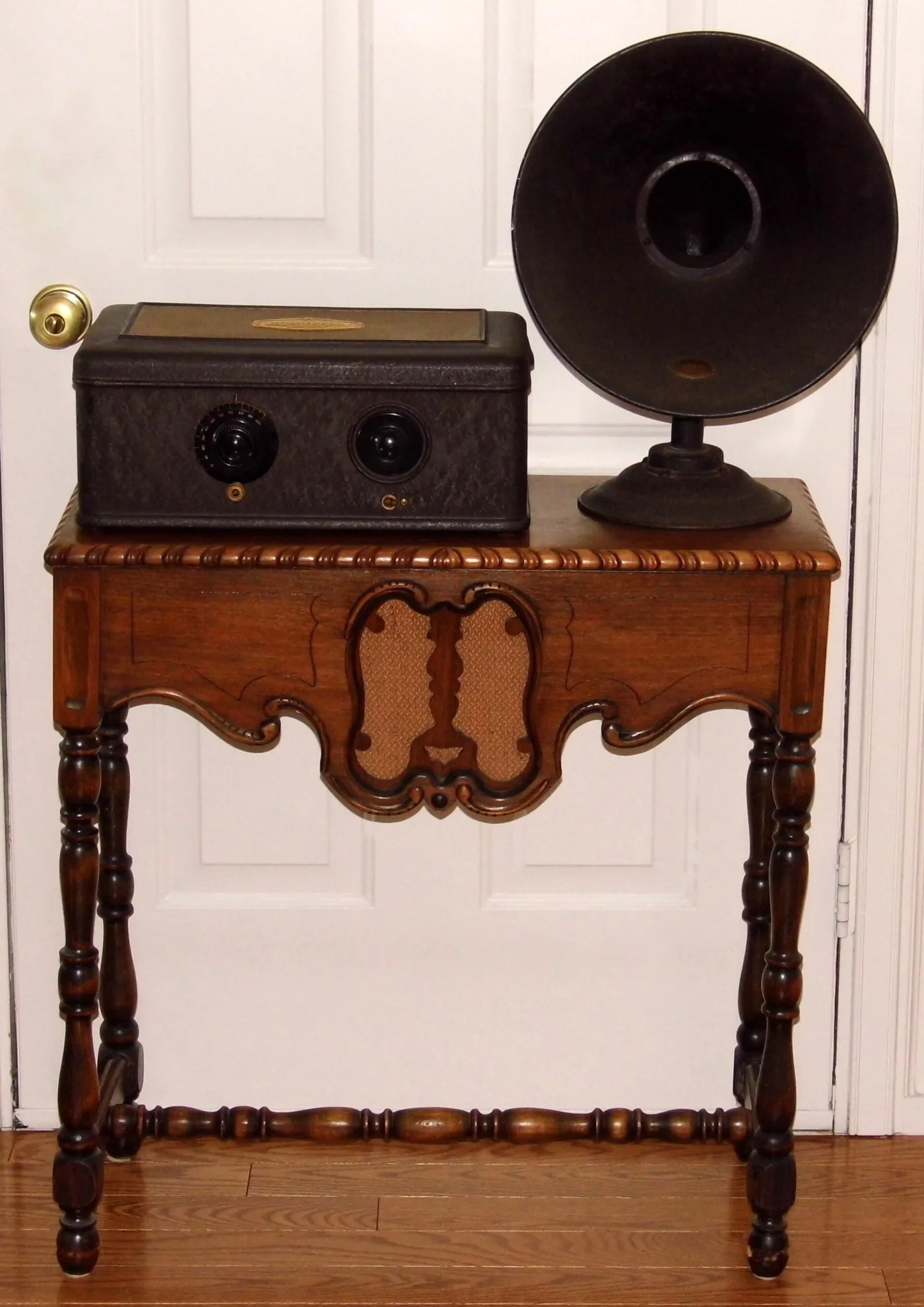 Black metal box radio with two controls standing on a wooden side table, beside a tall black horn speaker on a round pedestal base
