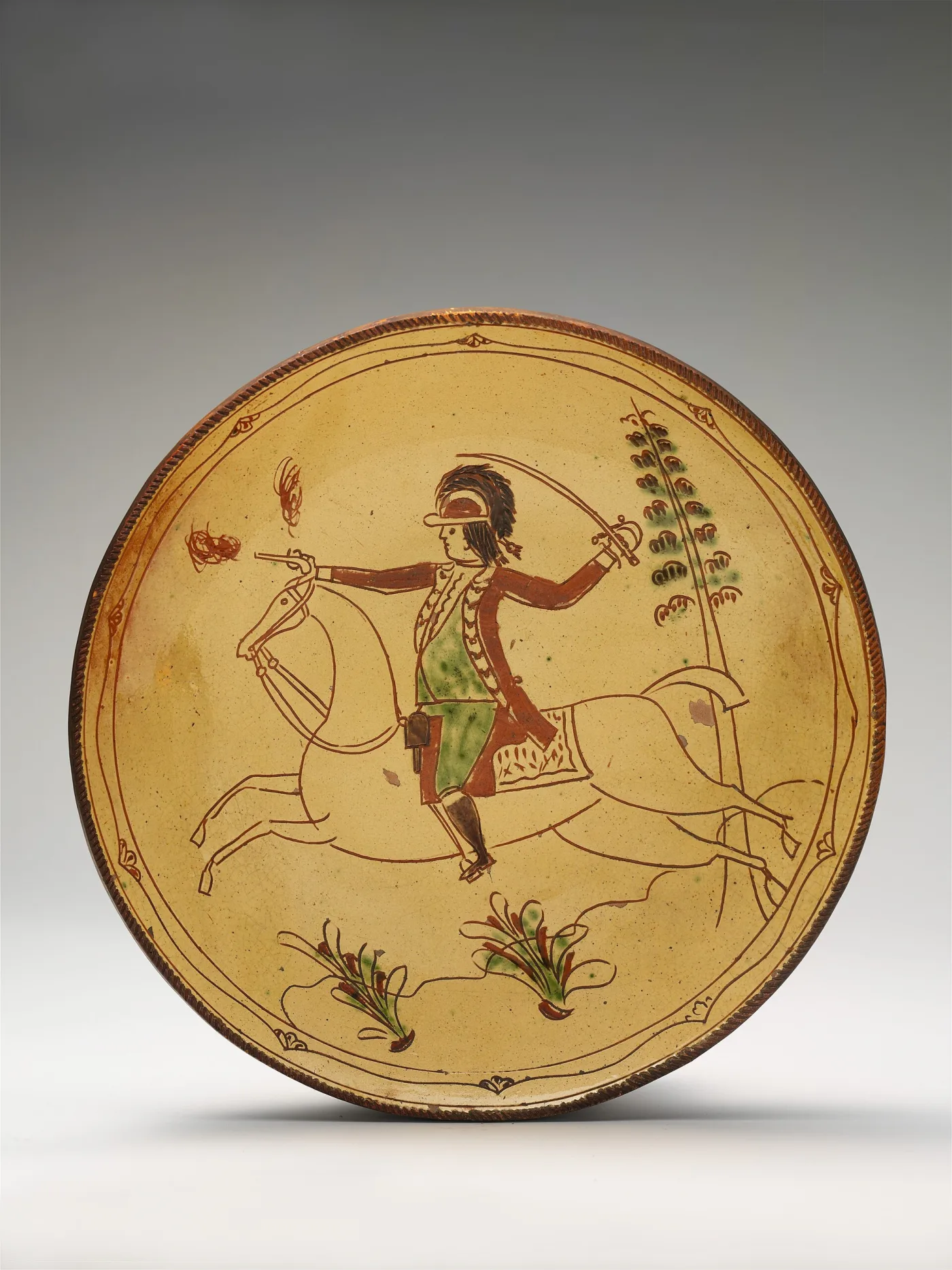 Buff-slipped redware plate incised with a mounted soldier brandishing a sabre, with green and red-brown accents and a coggled rim