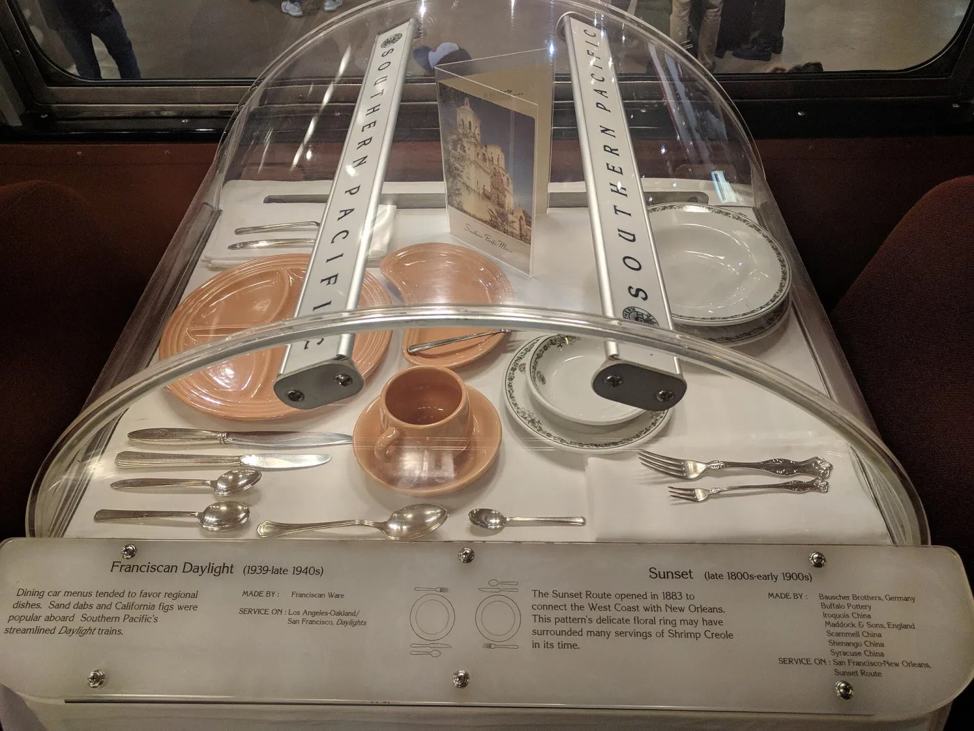 Museum case of Southern Pacific china, a coral-pink set beside a white service with a delicate floral ring, and an interpretive label naming seven potteries