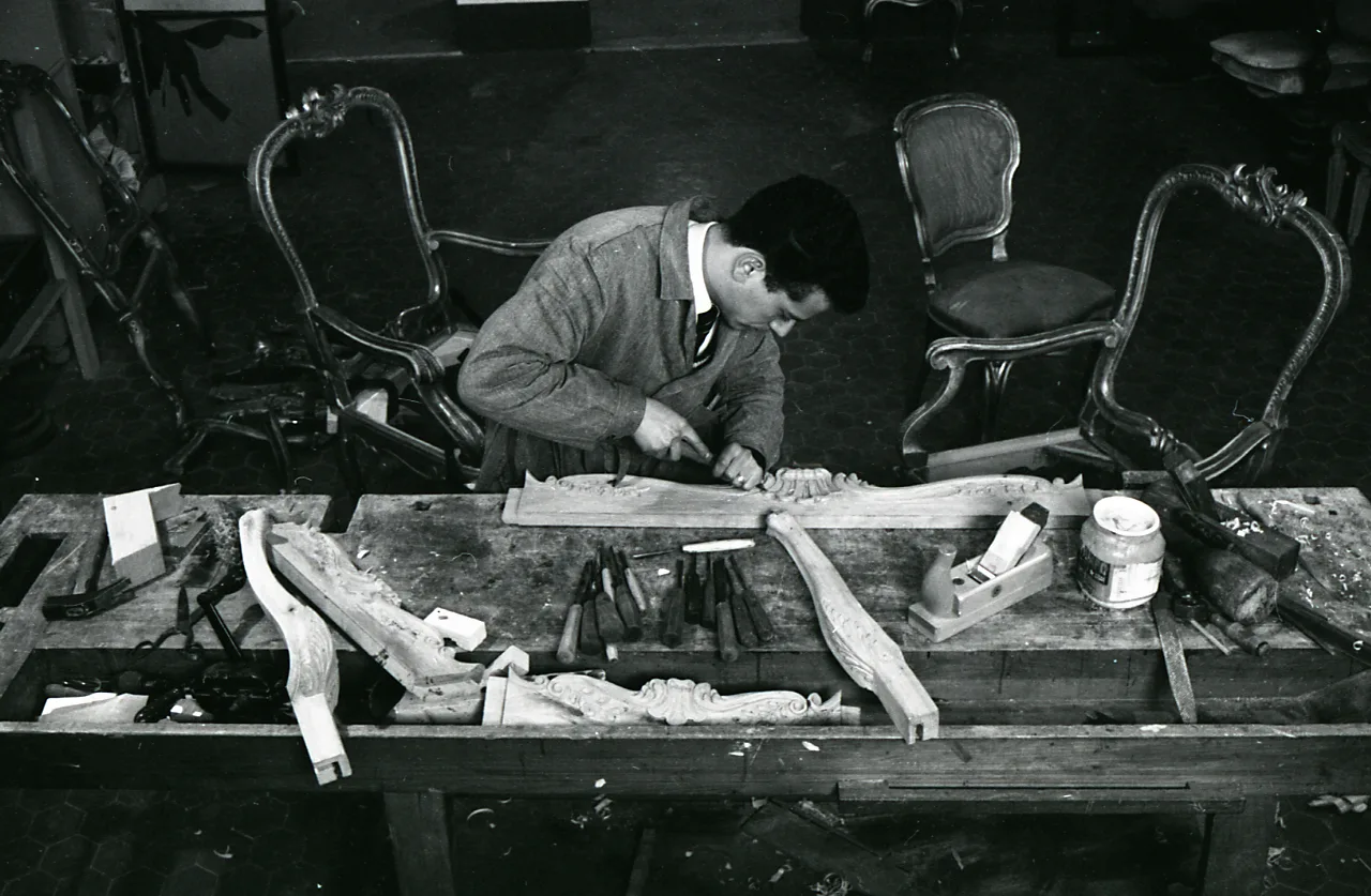Black and white photograph looking down on a workbench where a man carves a scrolled wooden crest, with gilded chair frames standing around him and carving gouges laid out in a row