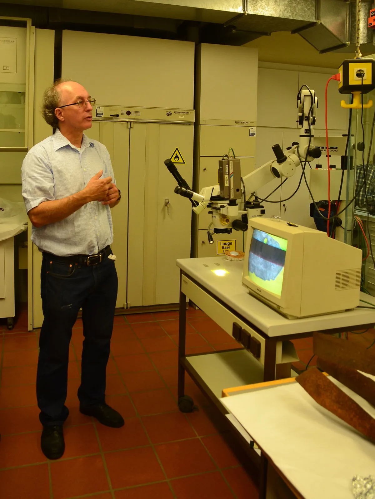 A conservator standing beside a stereo microscope fitted with a video head, its magnified view of a corroded object shown on a monitor, in a laboratory with fume cabinets and chemical warning signs