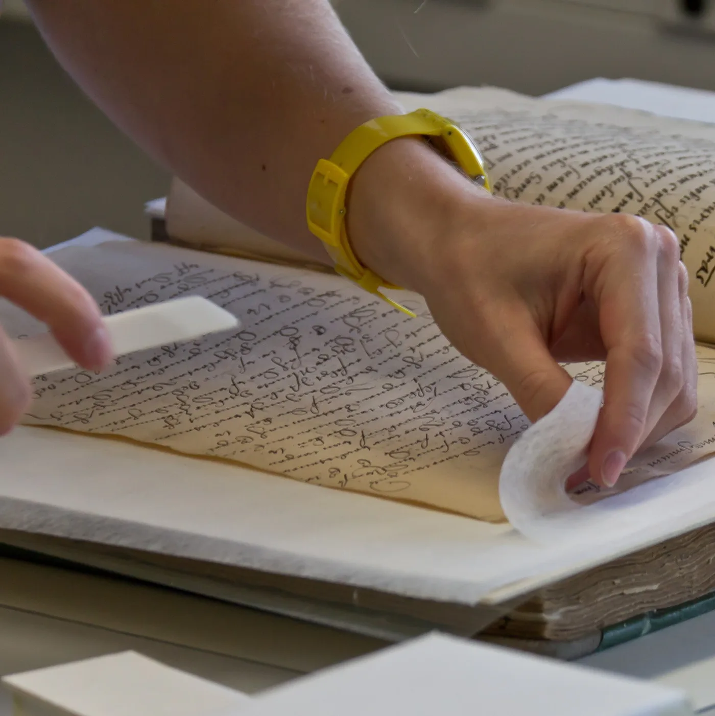 Two hands laying absorbent sheets between the leaves of an open manuscript volume written in brown iron-gall ink
