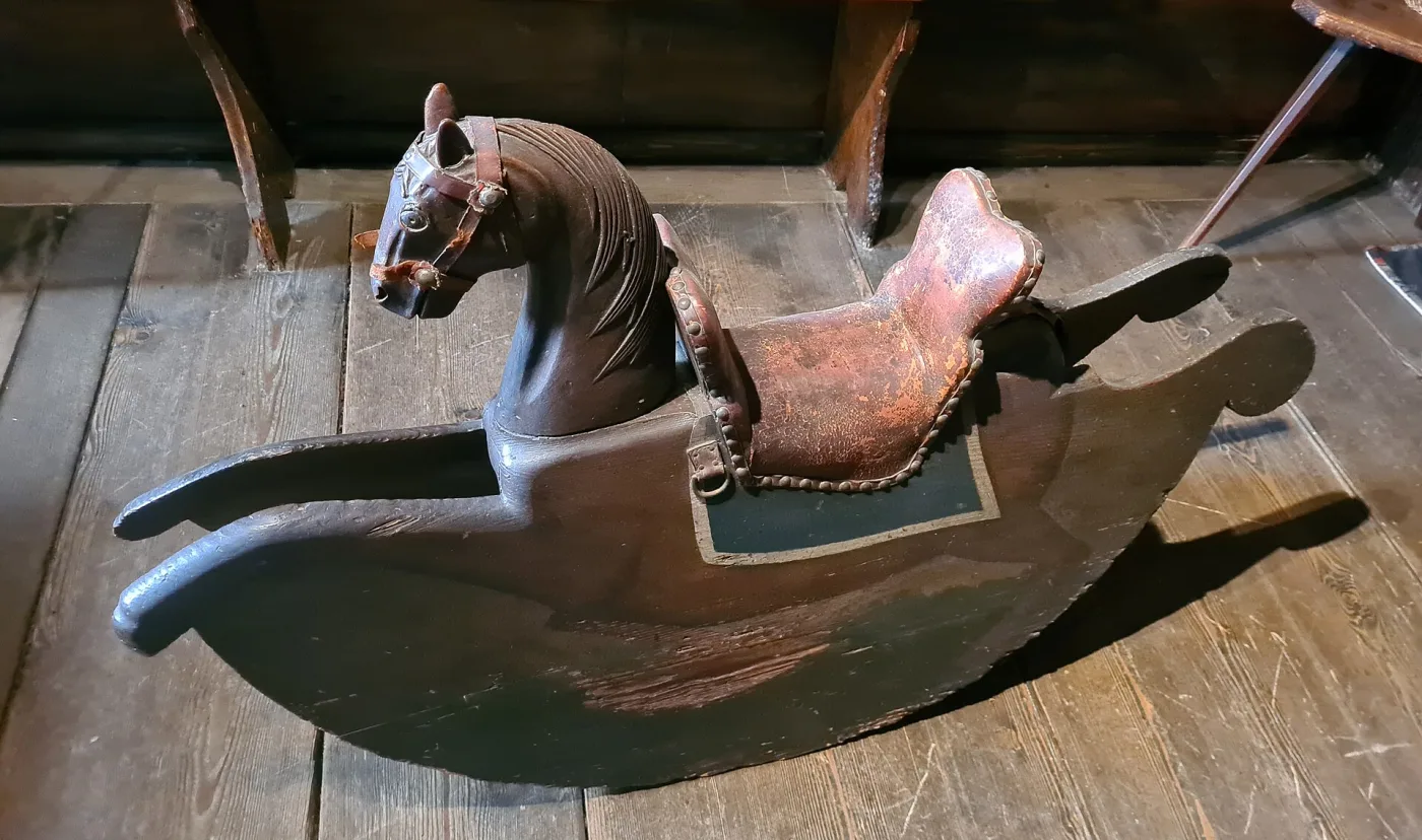 Dark carved wooden rocking horse with the mane cut into the neck as rows of locks, a studded red leather saddle over a green cloth, on solid shaped rockers