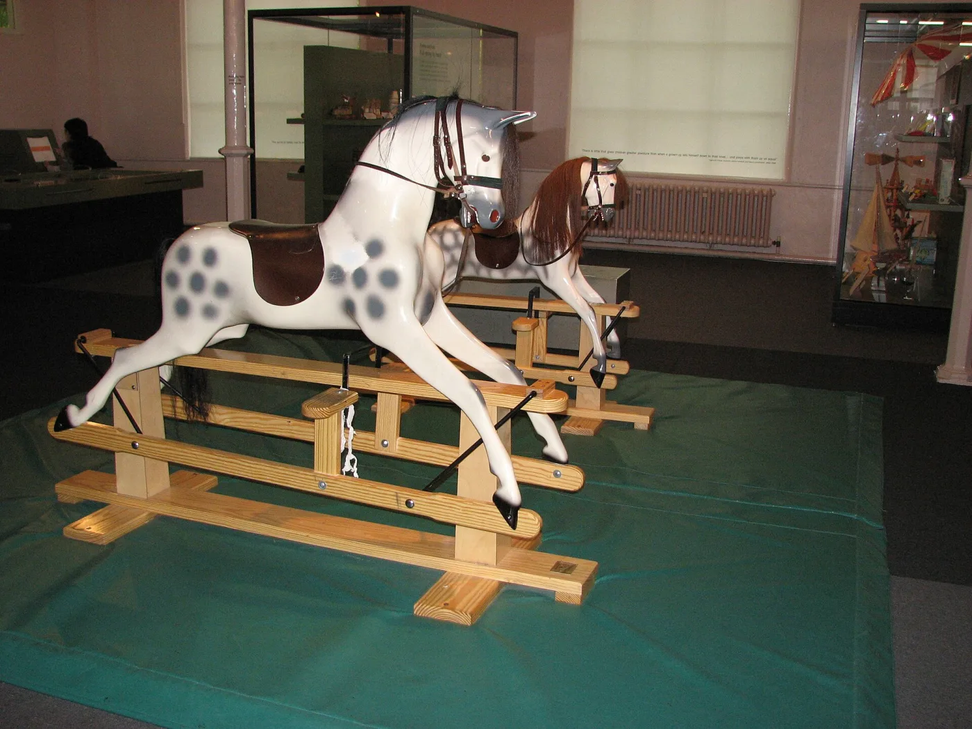 Dapple grey rocking horse hanging from black iron swing straps on a rectangular pine trestle stand, with a second horse behind it