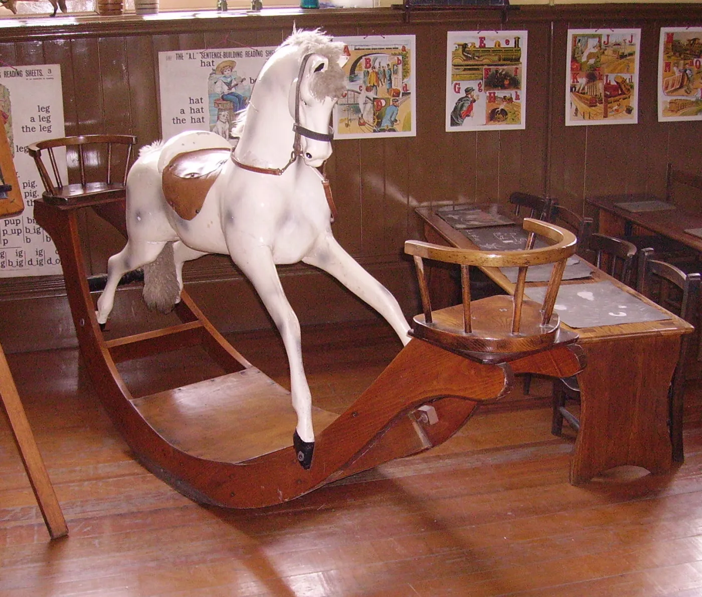 White dapple grey rocking horse with a horsehair mane and brown leather saddle mounted on shaped wooden bow rockers, in a museum schoolroom
