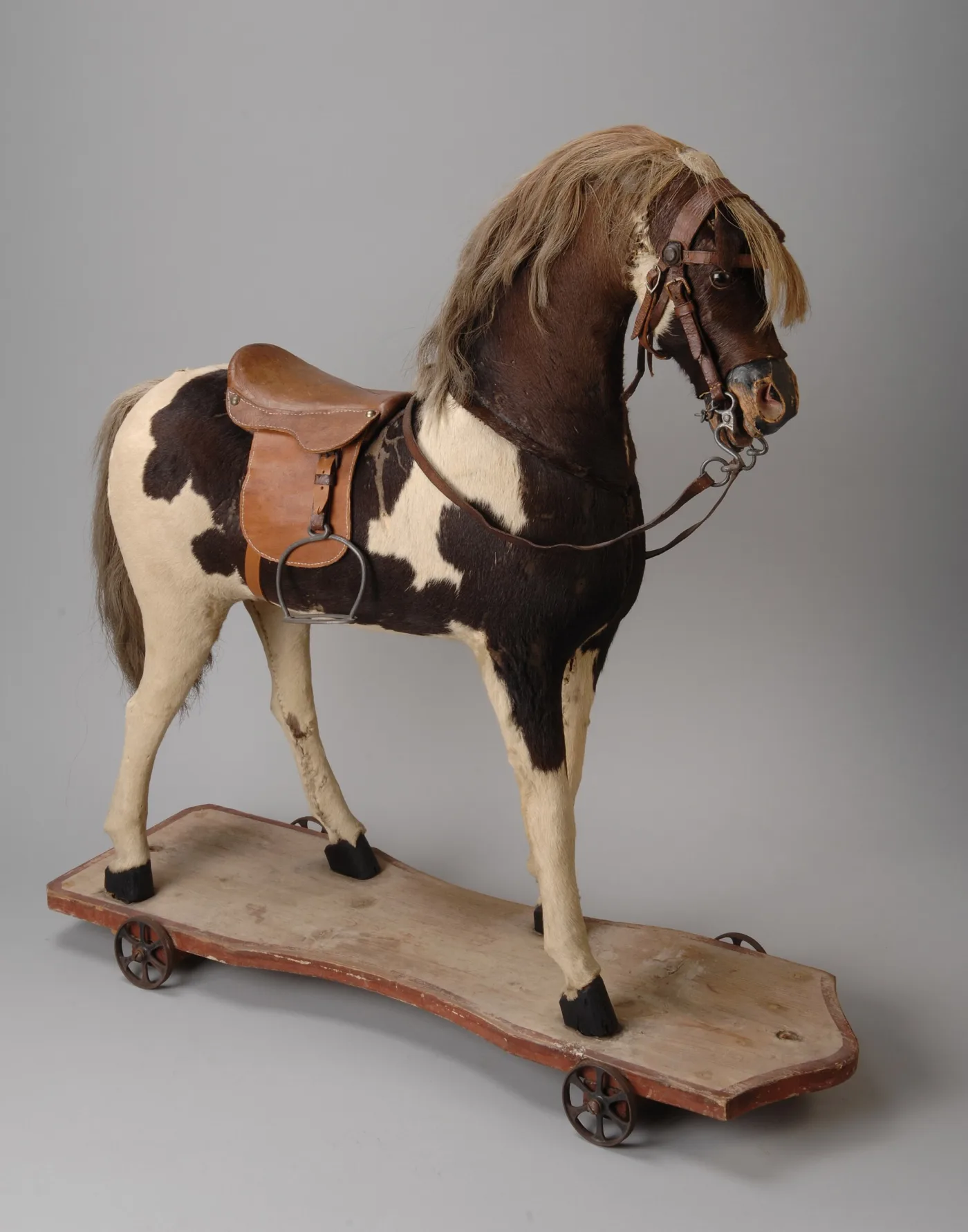 Brown and white hide-covered toy horse with a horsehair mane, leather saddle and metal stirrup, standing on a red-edged wooden platform with four cast iron wheels