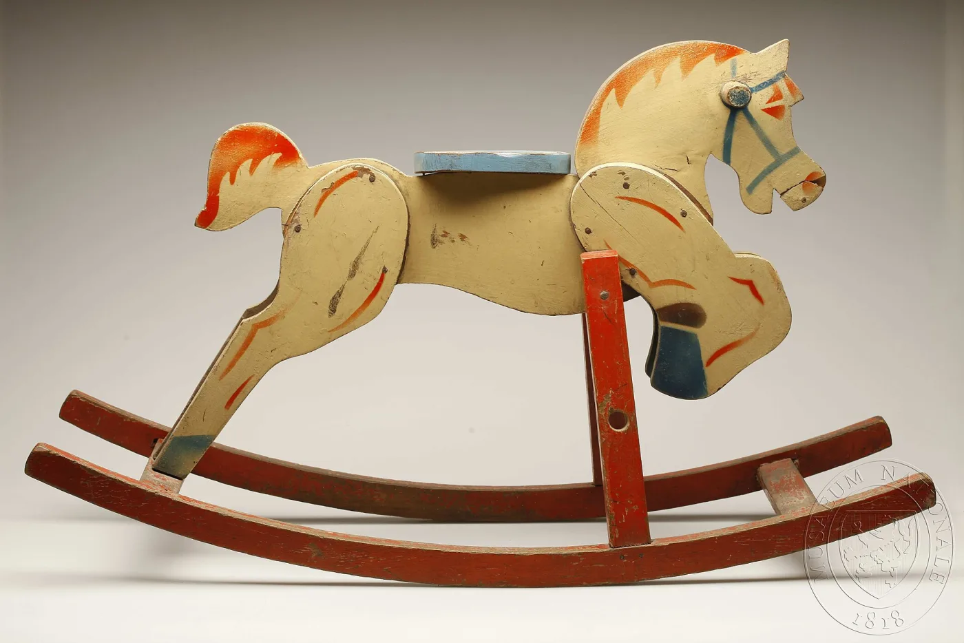 Cream-painted rocking horse cut from flat boards with orange and blue painted detail and a round blue seat, mounted on red-painted bow rockers