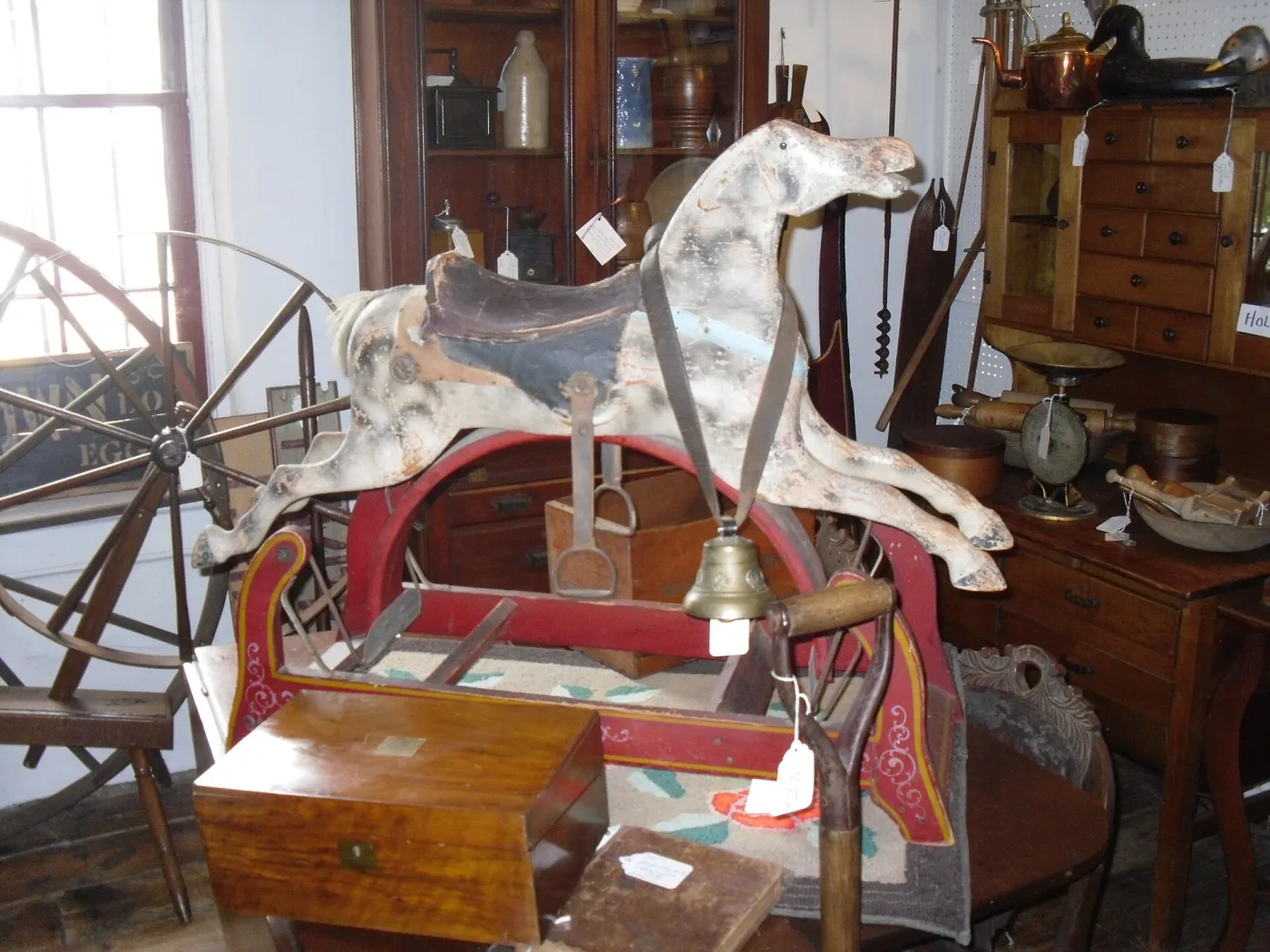 Worn white and grey rocking horse with paint rubbed through to bare wood, a dark leather saddle, stirrup and hanging brass bell, on a red painted rocking frame in a crowded antique shop