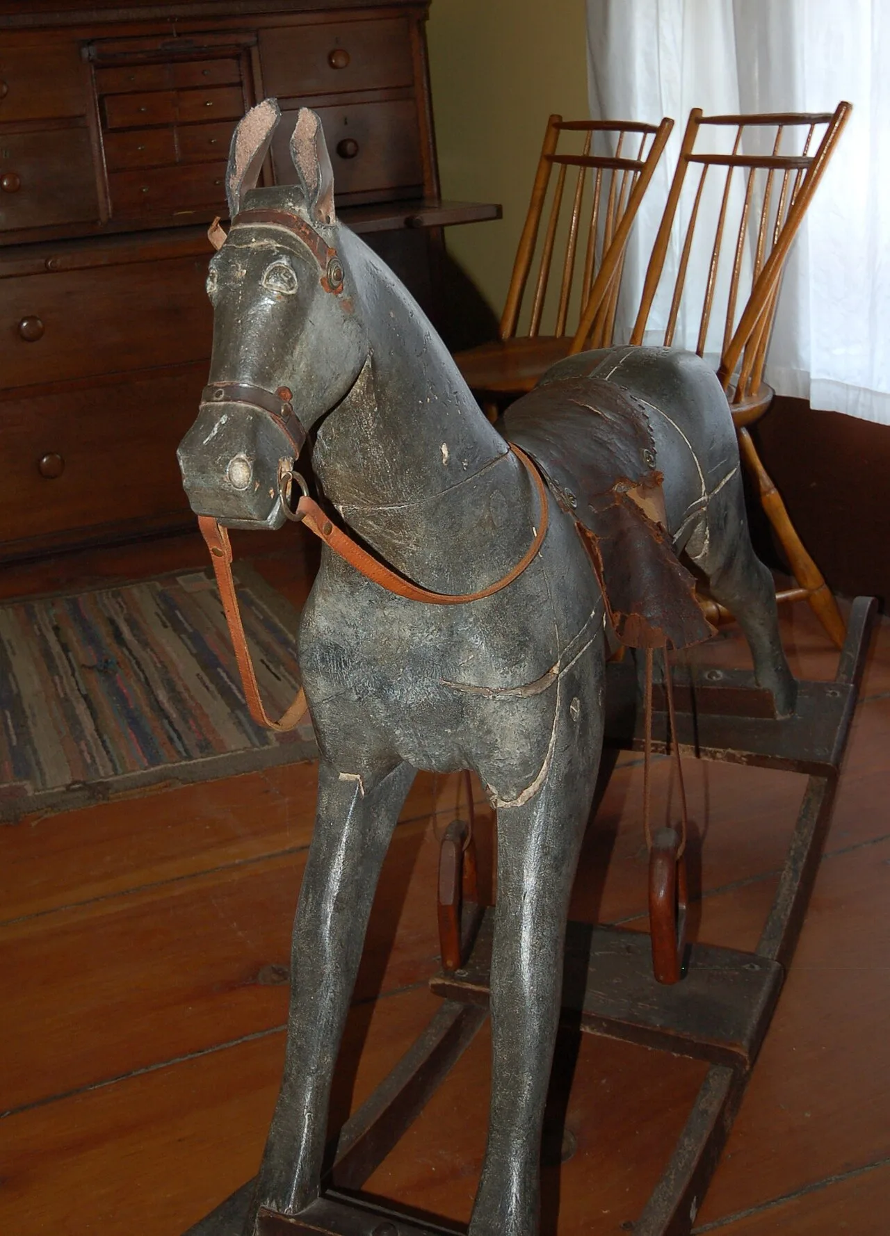 Grey painted carved wooden horse with worn paint, split leather saddle and leather bridle, standing on a dark wooden rocking frame in a period room