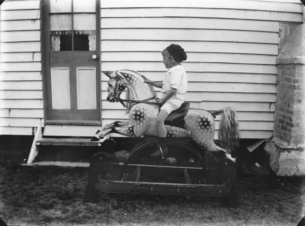 Black and white photograph of a boy in a cap seated on a large dapple grey rocking horse with a long horsehair tail, its curved rockers set inside a rectangular wooden base