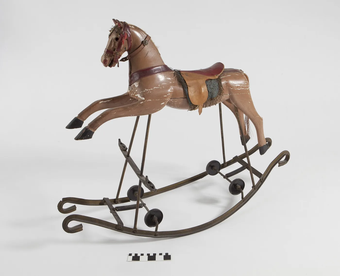 Pink-brown painted wooden horse with a leather saddle and fringed cloth, mounted on curved wrought iron rockers fitted with coil springs and small folding wheels, with a photographic scale bar below