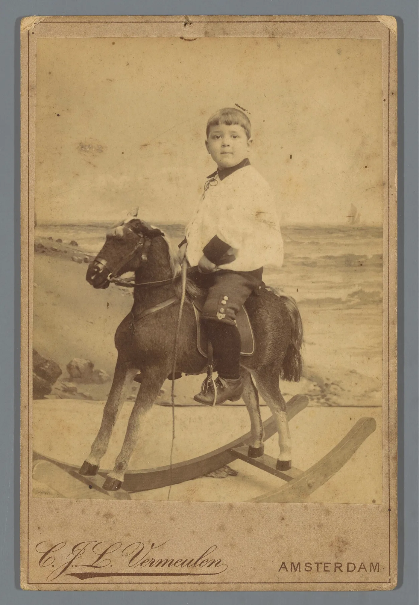 Sepia cabinet photograph of a small boy in a white blouse seated on a hide-covered rocking horse standing on plain wooden bow rockers