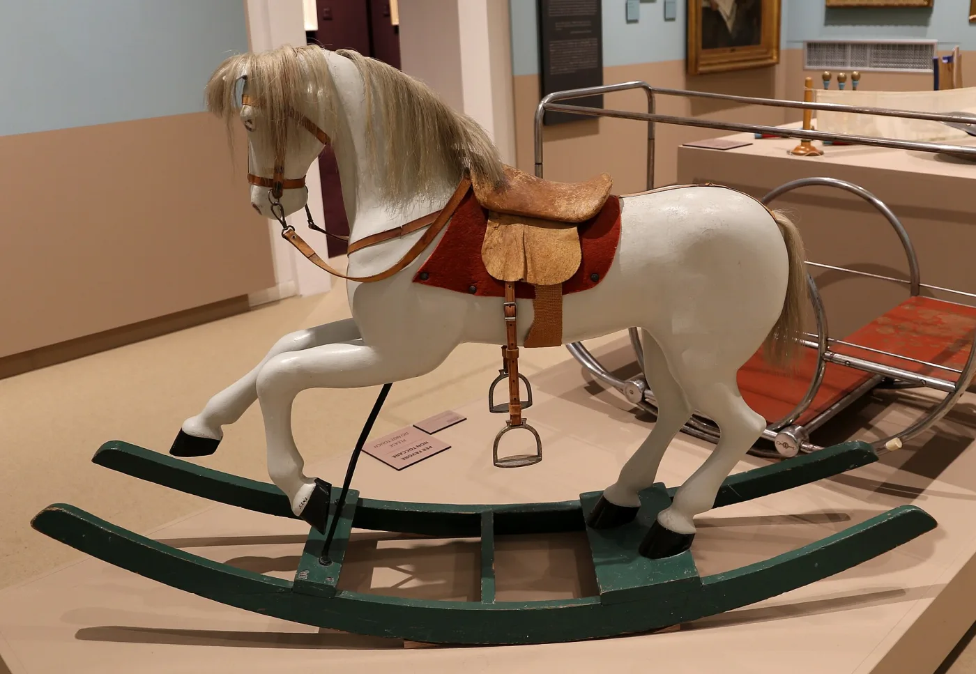 Pale grey painted rocking horse with a blond horsehair mane and tail, leather saddle and metal stirrups, standing on dark green curved bow rockers in a museum display
