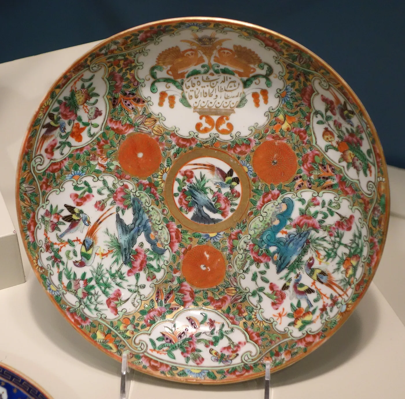 Chinese export dish of about 1865 with rim panels containing only birds, butterflies, flowers and fruit, a bird and peony central roundel, four plain orange discs, and a Persian inscription cartouche at the top