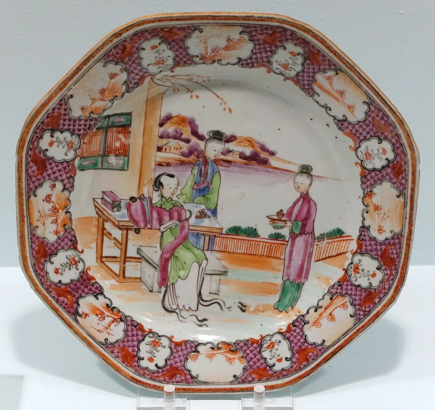 Octagonal Chinese export plate of about 1770 to 1780 with one large scene of a seated woman at a table attended by servants filling the whole well, inside a crimson diaper border with white floral reserves