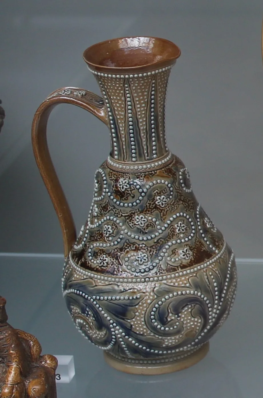 Tall stoneware jug with a strap handle, its neck covered in a dense pattern of small applied beads and its body incised with bold scrolling foliage in blue on buff