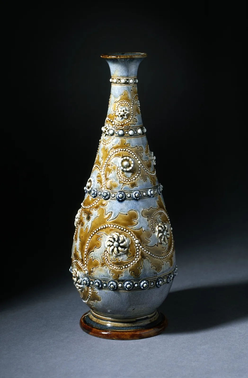 Slender bottle-shaped vase with a long neck, glazed in pale blue-grey and ochre and covered with bands of applied beading and small white rosettes between incised borders