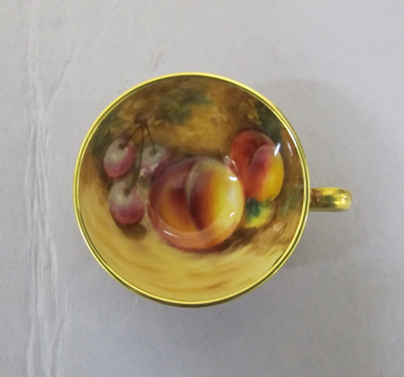 Interior of a small porcelain cup painted with a peach, plums and cherries lying on a mossy bank, with a gilded rim and a painted signature at the lower right