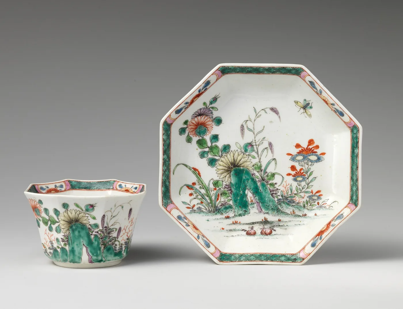 Octagonal handleless teabowl beside a matching octagonal saucer, painted with green rockwork, flowers and a butterfly