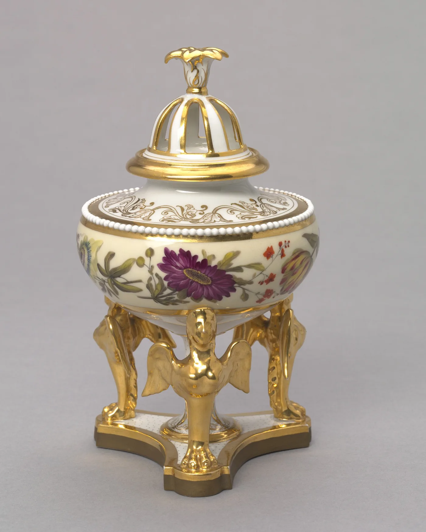 Covered urn painted with flowers on a cream ground, with a pierced gilt dome cover and three gilt winged-figure supports on a triangular base