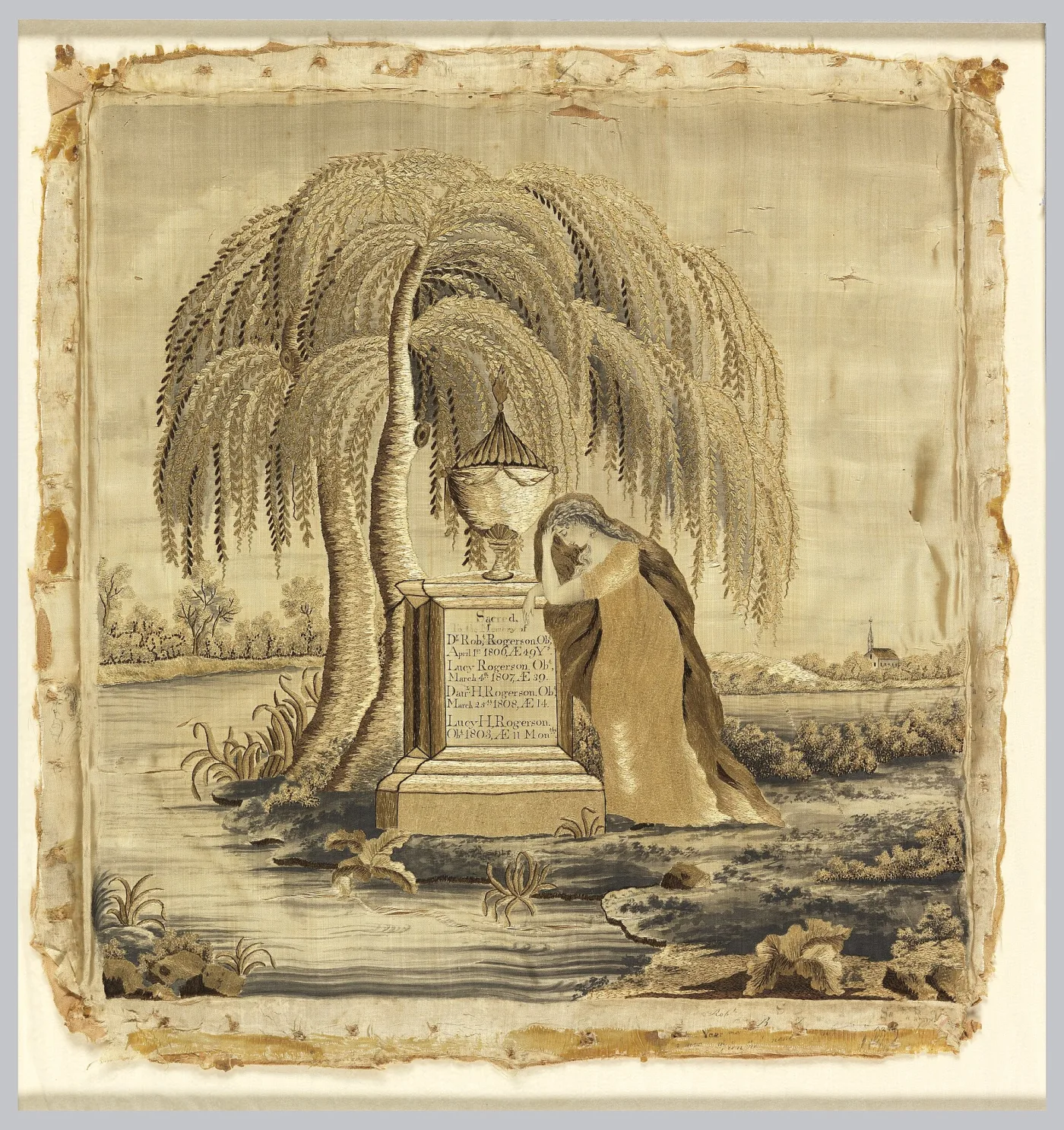 Silk embroidered picture of a woman in a long gown leaning on a tomb topped by an urn beneath a large weeping willow, with a river, a distant church and an inscribed tomb face