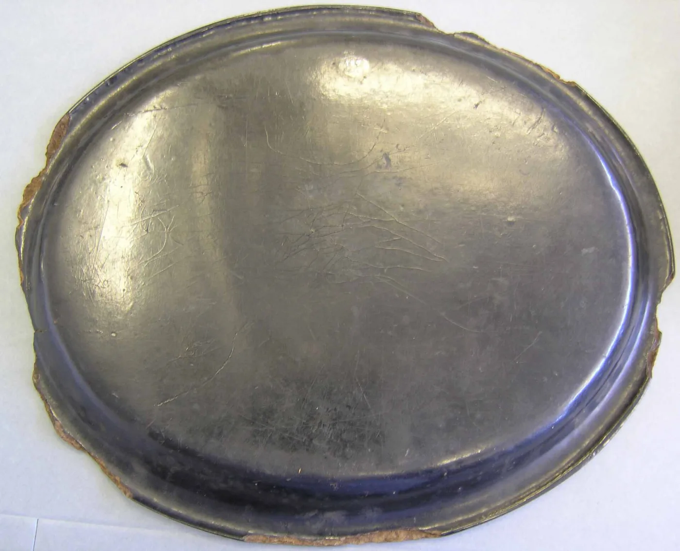 Underside of an oval black tray photographed flat, the lacquer surface scratched and dulled, with the rim broken away all round to expose a rough brown fibrous layer