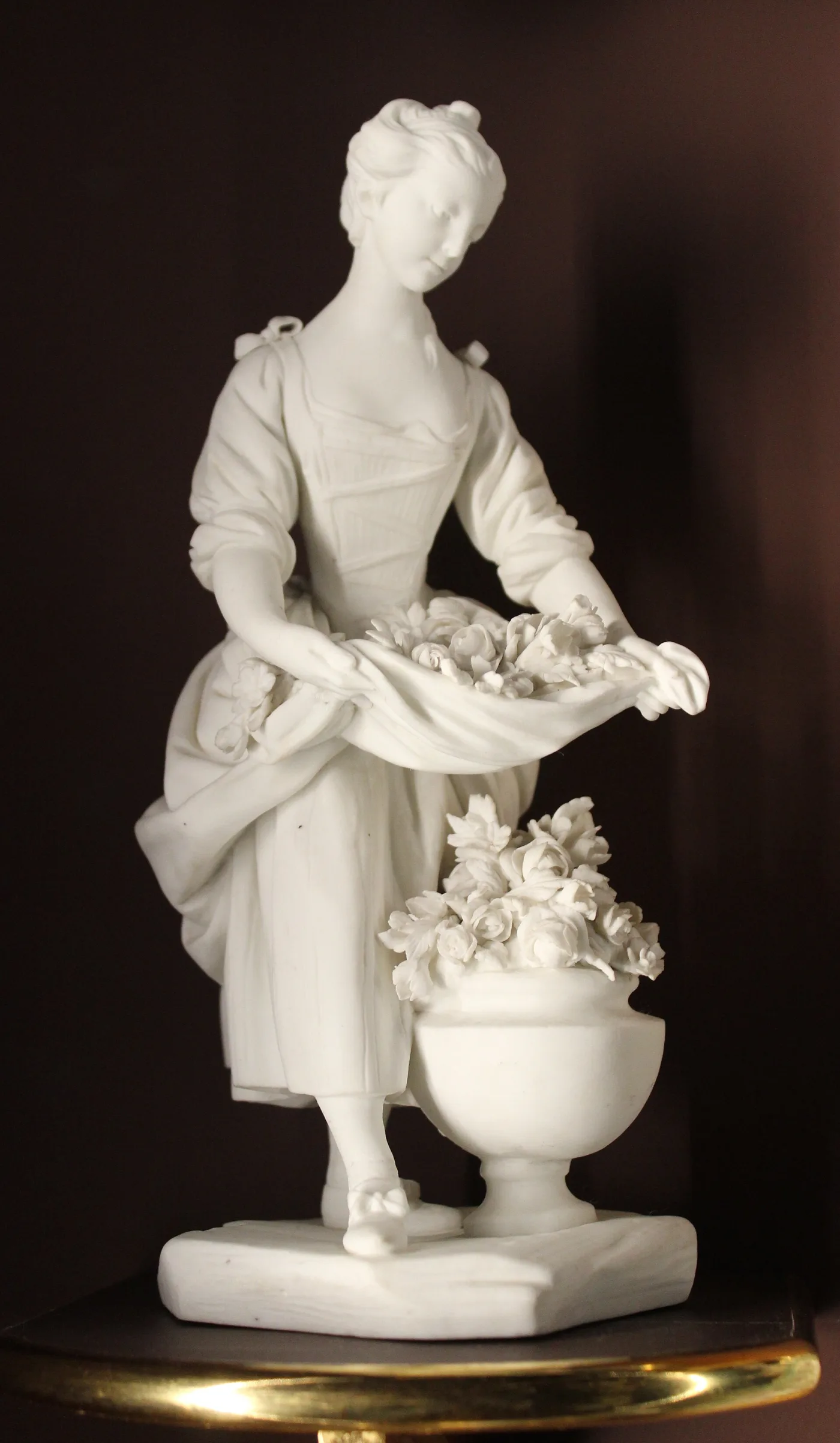 Unglazed white porcelain figure of a young woman holding flowers gathered in her apron, standing beside an urn of modelled roses