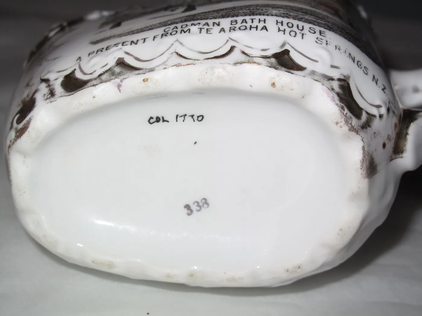 The underside of the same white shaving mug, showing a curved printed inscription reading present from Te Aroha Hot Springs and a hand-written accession number