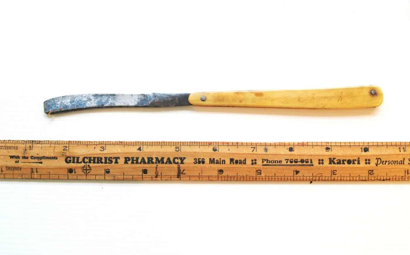 The same early straight razor photographed alone on a ruler, its bone handle pale and its steel blade covered in blue-grey corrosion from heel to point