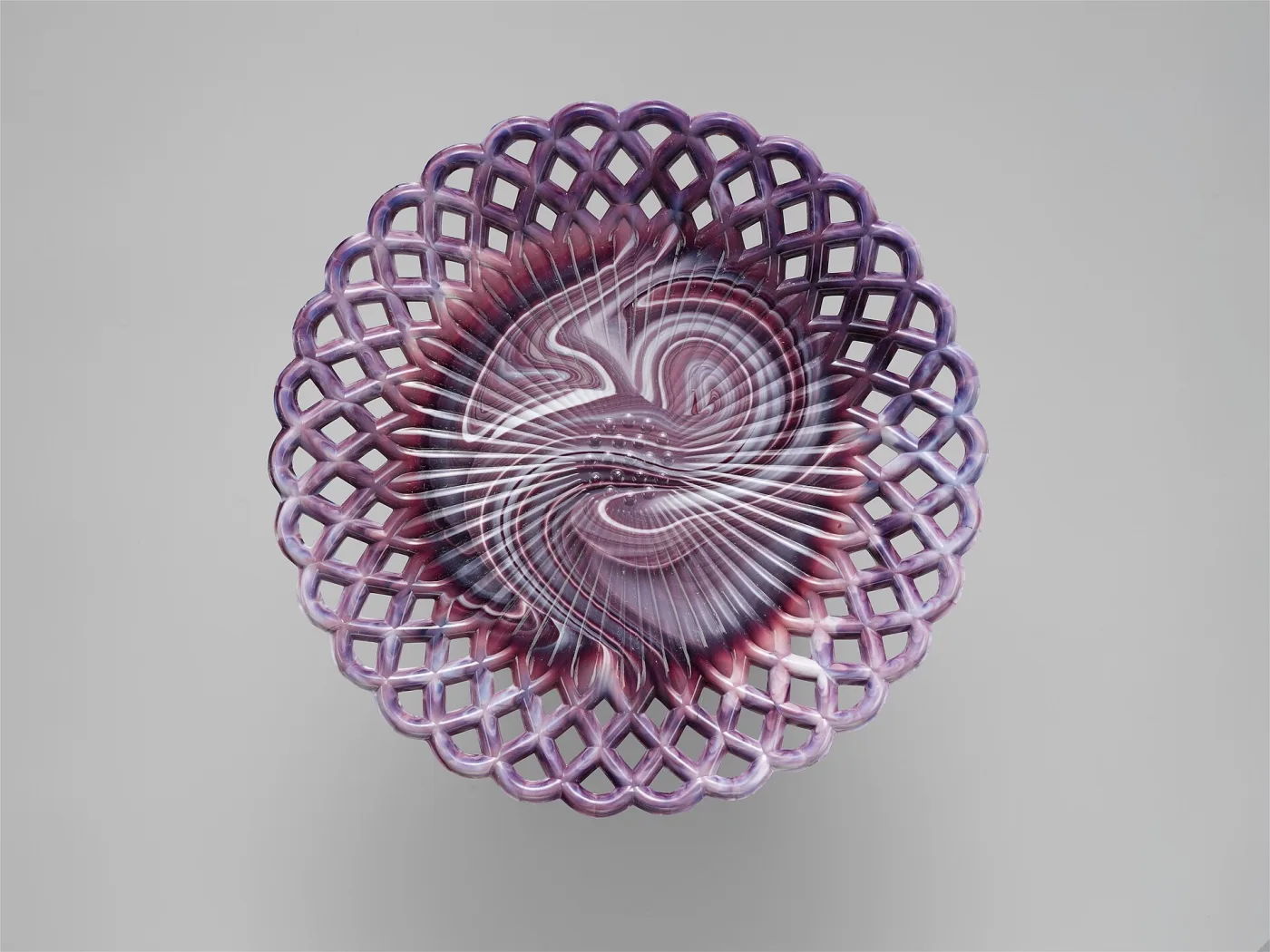 Circular purple and white marbled glass plate photographed from directly above, its rim an open lattice of interlocking loops, the centre swirled with white streaks radiating outward