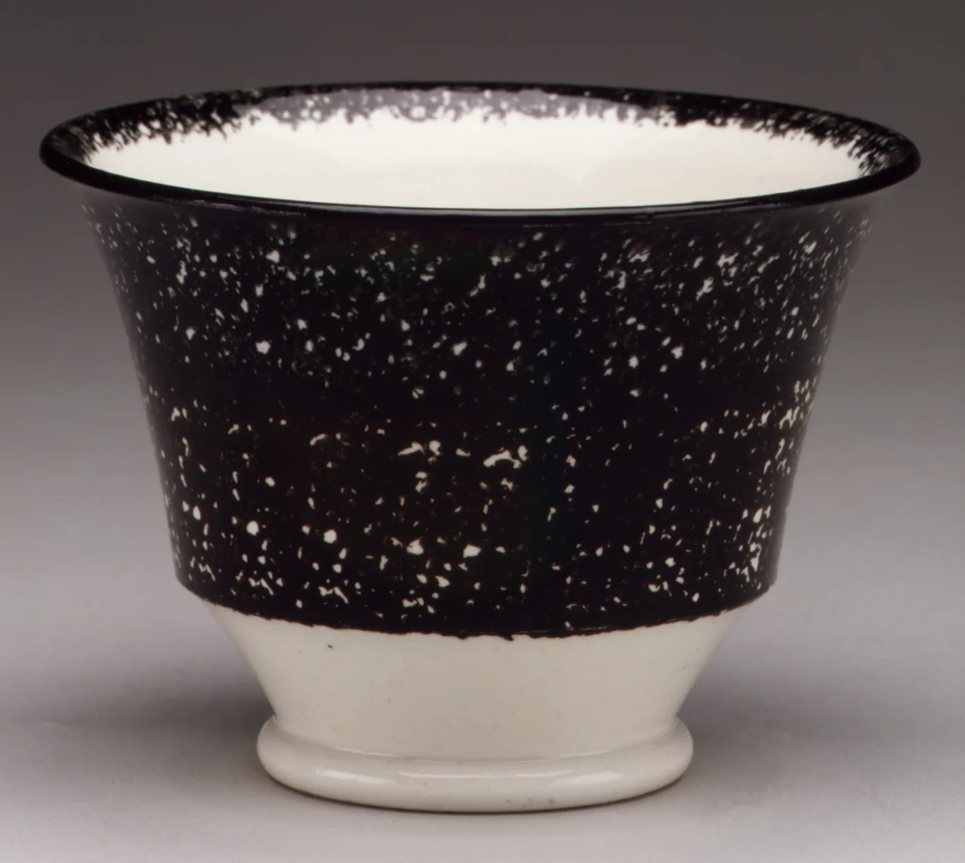 A handleless cup covered in dense black sponging over its upper body, left plain white towards the foot, the interior white with a sponged rim