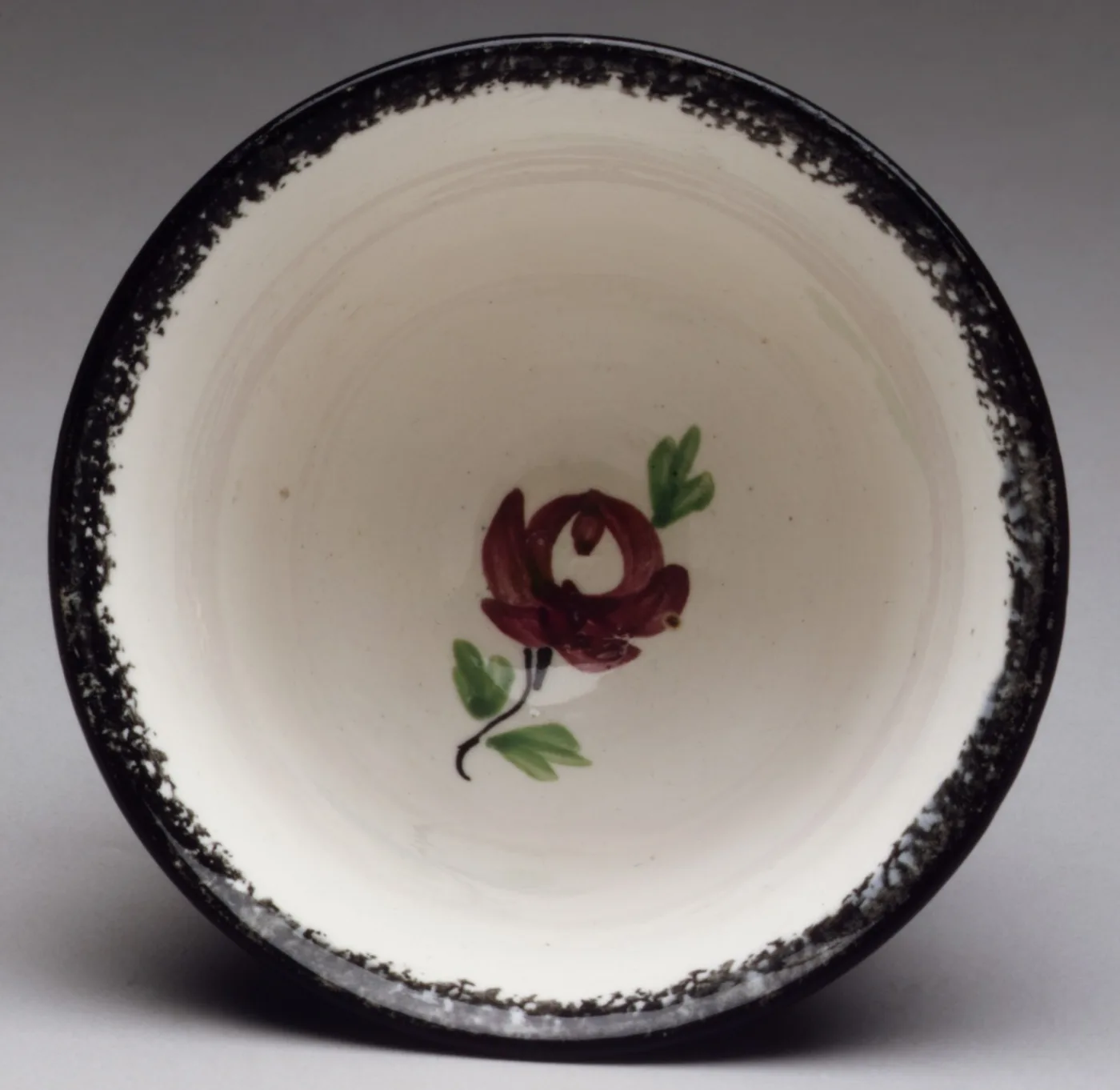 The inside of a cup, white with a black sponged band around the rim and a small hand-painted red rose with green leaves in the centre