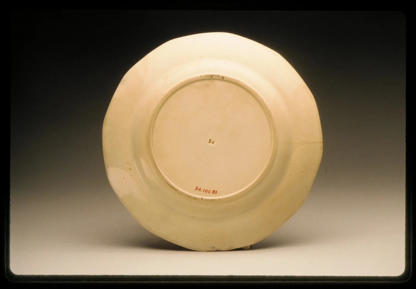 The plain cream-coloured underside of a plate, showing a small impressed device near the centre and a museum accession number painted in red