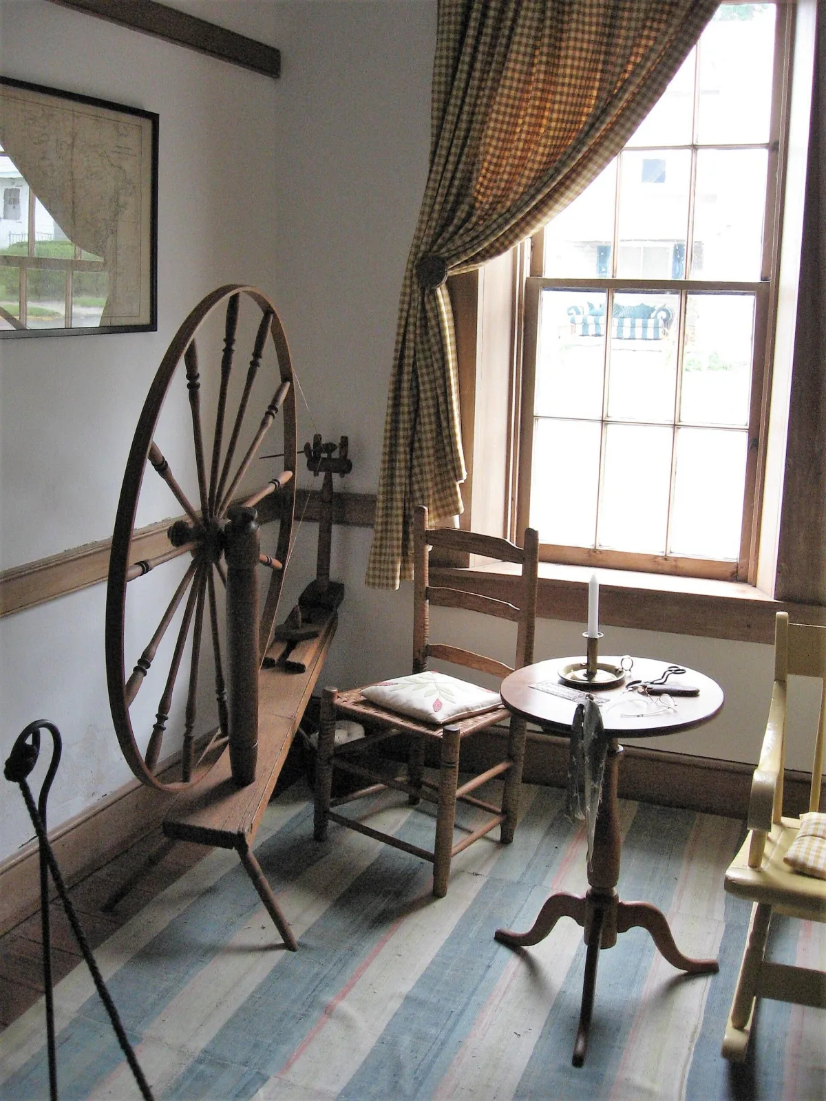 Large wooden spinning wheel with a tall spoked drive wheel at one end of a long angled bench standing on three splayed legs, with a small spindle head at the low end, in a plain painted room