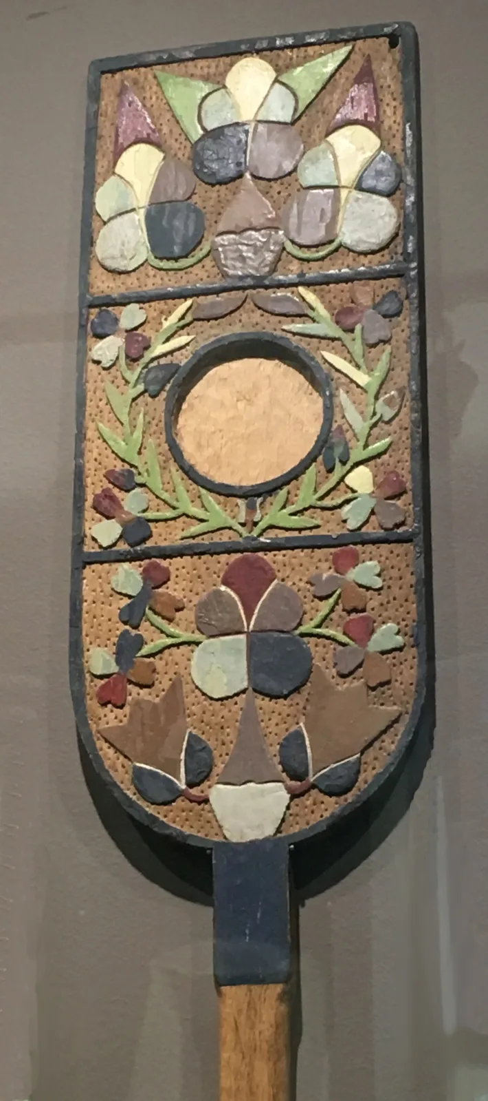 Close view of a distaff blade carved and painted with flower panels on a stippled ground, divided into three fields, with an empty circular recess ringed in black at its centre