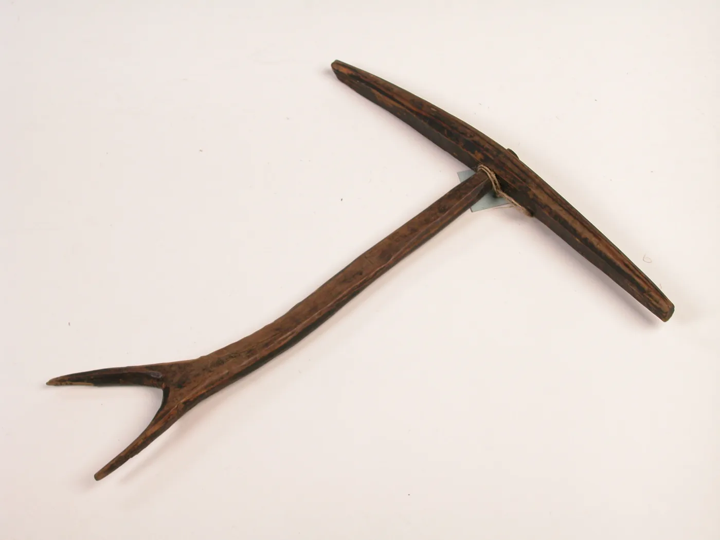 Dark brown wooden tool photographed on white, a central shaft with a crossbar at each end set at right angles to one another, one end forked, with a small museum tag tied to it