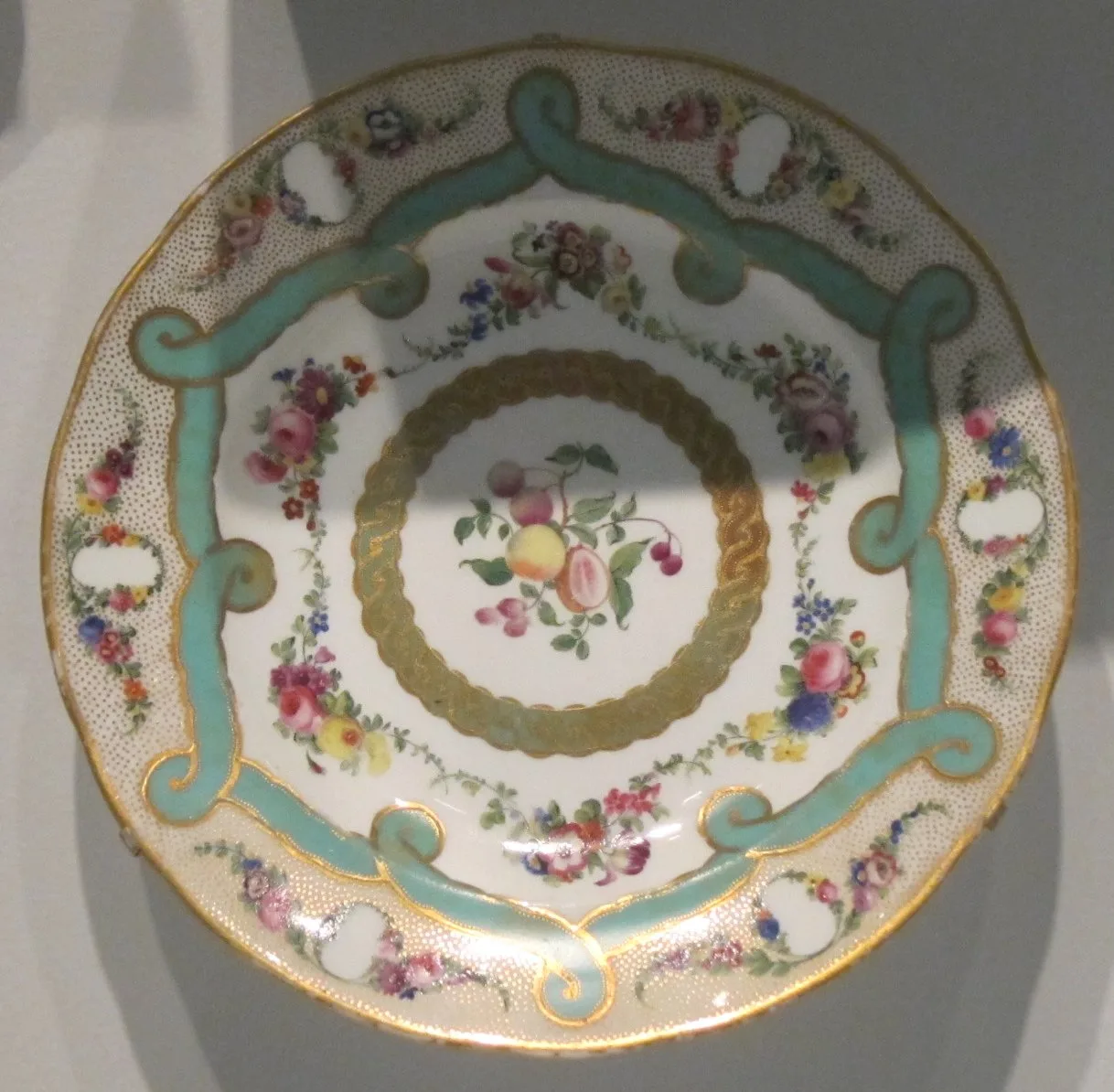 Porcelain plate of 1846 with a pale green scrolling band and gilt stippling round the rim, enclosing reserved panels and sprays of painted roses and fruit around a small gilt-ringed centre
