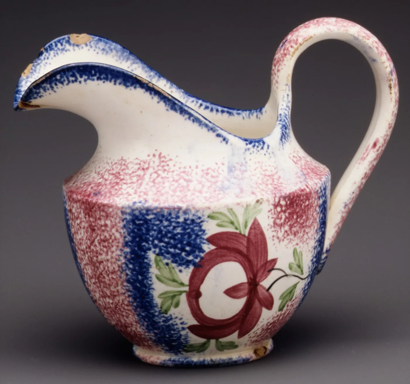 Panelled pitcher of about 1800 to 1830 with vertical bands of red and blue spatter, a hand-painted red rose with green leaves in a white reserve, and blue spatter running along the rim, spout and handle