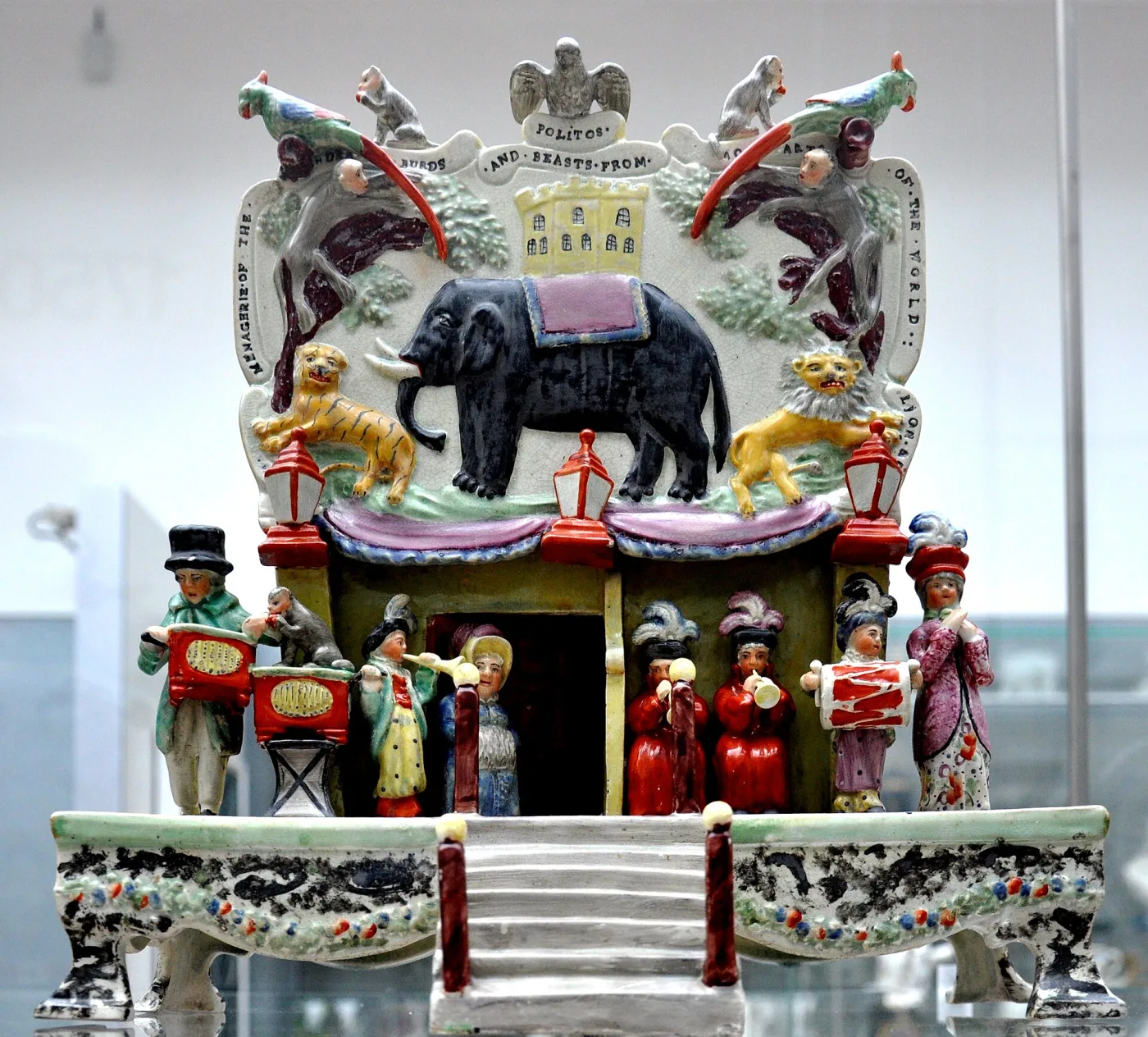 Pottery model of a fairground menagerie: a lettered plaque with an elephant, tiger and lion in relief above a booth, with a band of musicians below, all on a table base with scrolled legs