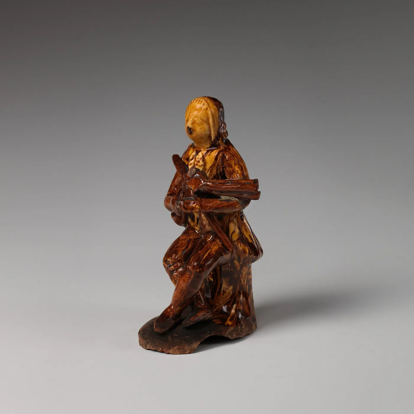 Small pottery figure of a seated bagpiper covered in streaked brown and ochre glazes over a cream body